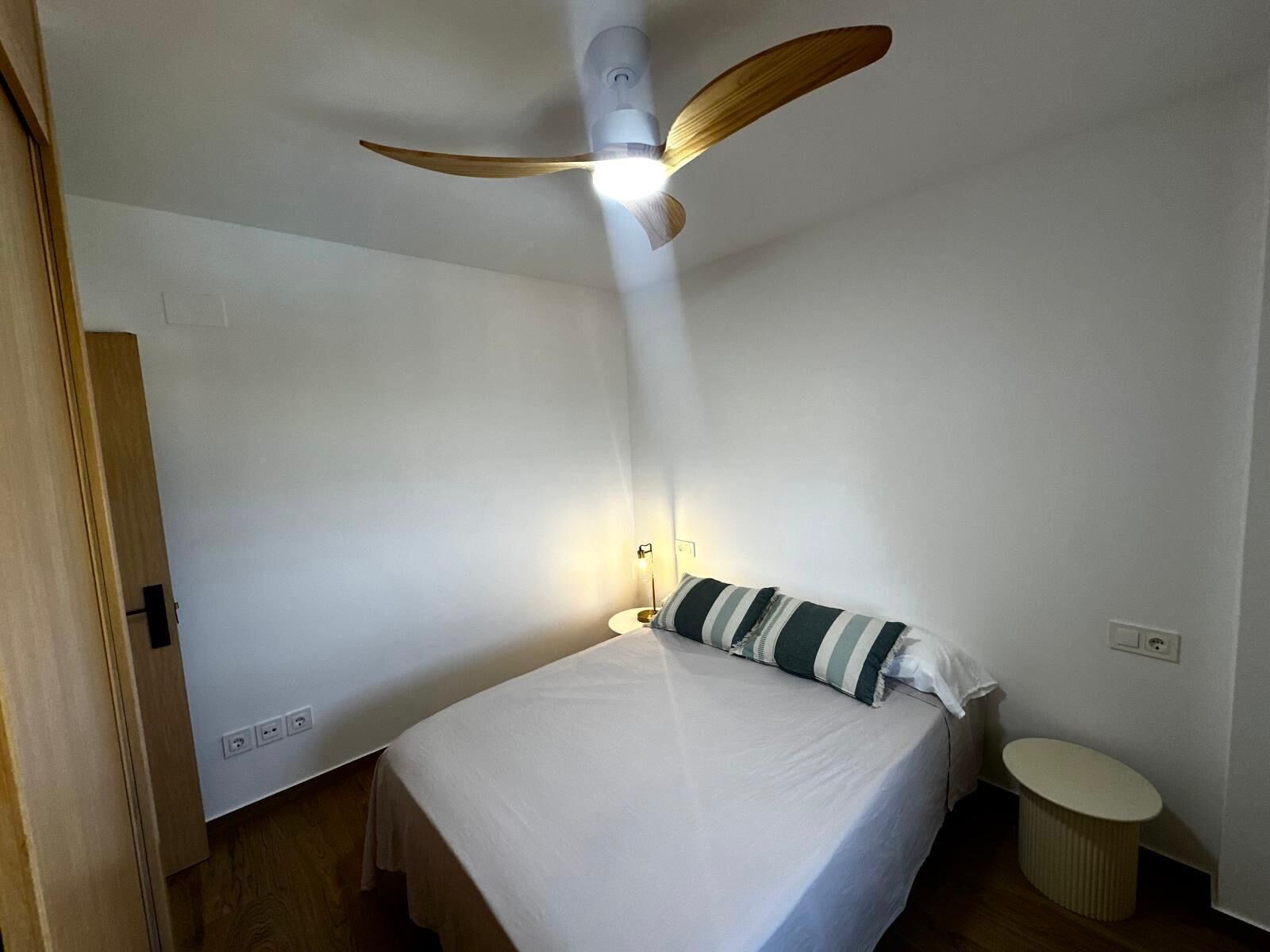Una habitación pequeña y limpia con una cama individual, un ventilador de techo, una lámpara de noche y una mesita redonda.