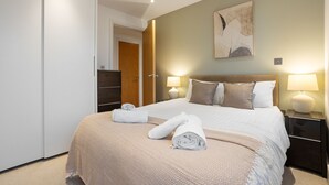 Un lit bien fait, recouvert d’une couvre-lit à motifs beige et blanc, avec deux oreillers et deux serviettes enroulées posées sur le lit. Une table de chevet avec une lampe se trouve à côté du lit, et une œuvre d’art est accrochée au mur au-dessus du lit.