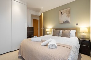 A neatly made bed with a beige and white patterned bedspread, two pillows, and two rolled towels on the bed. There is a nightstand with a lamp and a wall-mounted artwork above the bed. - 25% OFF! | City Wide Views | 3B Canary Wharf (Greater London)