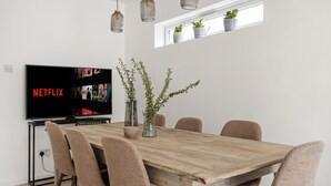 Zona pranzo con tavolo in legno, sei sedie, una TV che mostra Netflix e due piante in vaso su uno scaffale.