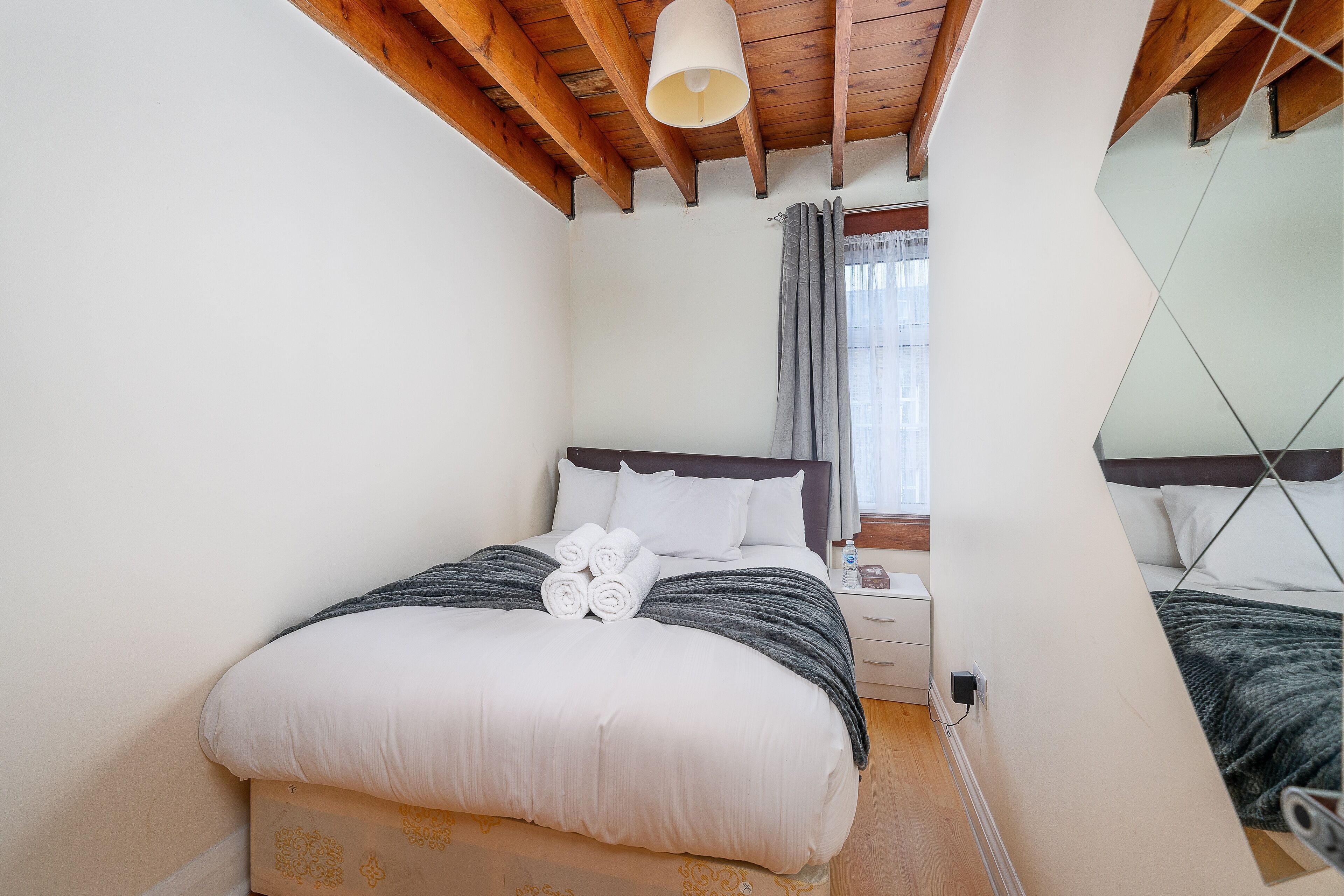 A neatly made bed with white and grey linens, a bedside table with a lamp, and a window with curtains.