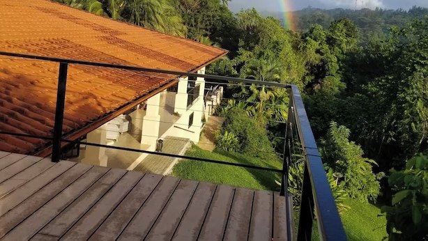 Eine Holzterrasse mit Glasgeländer, die auf eine üppige Landschaft und einen Regenbogen blickt.
