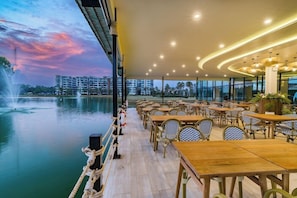 Un restaurante al aire libre con mesas y sillas, con vista a un cuerpo de agua y un cielo al atardecer.