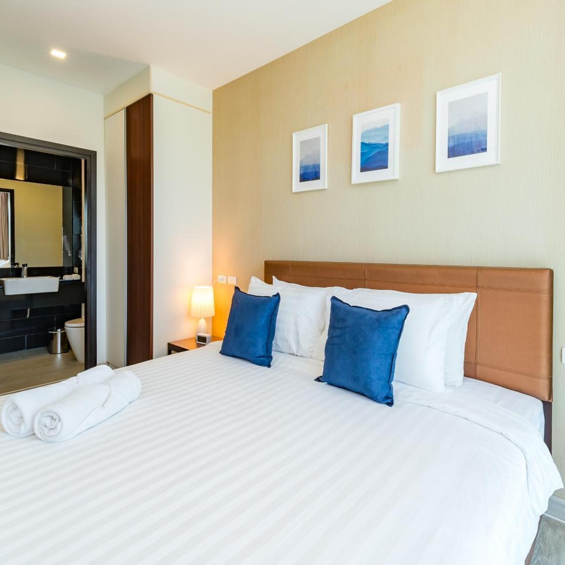 A hotel room with a large bed, a nightstand with a lamp, a bathroom with a mirror, and three framed pictures on the wall.