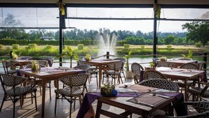 Un espace extérieur aménagé pour les repas, avec des tables et des chaises, un élément décoratif aquatique et une vue sur un plan d’eau et la végétation environnante.