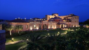 A large, multi-story building with a well-lit exterior, featuring a mix of stone and stucco facades, and a landscaped garden with palm trees. - Juna Mahal Jungle Resort Ranthambore (Sawai Madhopur)