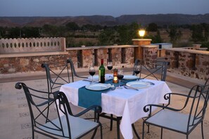 Mesa para cuatro personas dispuesta con copas de vino, una botella y un farol en una terraza con pared de piedra y vistas a colinas.