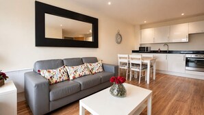 A modern living room with a grey sofa, a white coffee table with a vase of red roses, a dining area with white chairs, and a kitchen with white cabinets and appliances. - City View Apartment w/ Balcony & Gym Access (Manchester)