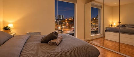 Un dormitorio moderno con una cama grande, una mesita de noche, un espejo y vista a un paisaje urbano al atardecer.
