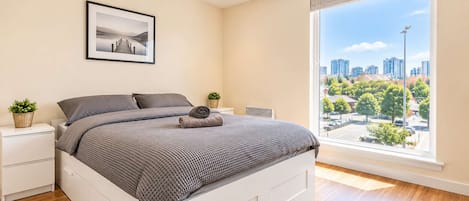 Een slaapkamer met een bed, nachtkastjes, een raam met uitzicht over de stad en een ingelijst beeld aan de muur.
