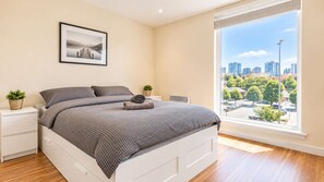 Un dormitorio con cama, mesitas de noche, una ventana con vista a la ciudad y un cuadro en la pared.