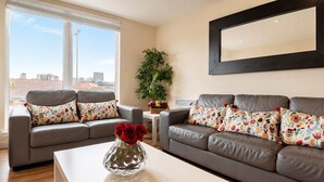 A living room with two grey sofas, colorful floral cushions, a white coffee table, and a vase with red roses. - City View Apartment w/ Balcony & Gym Access (Manchester)