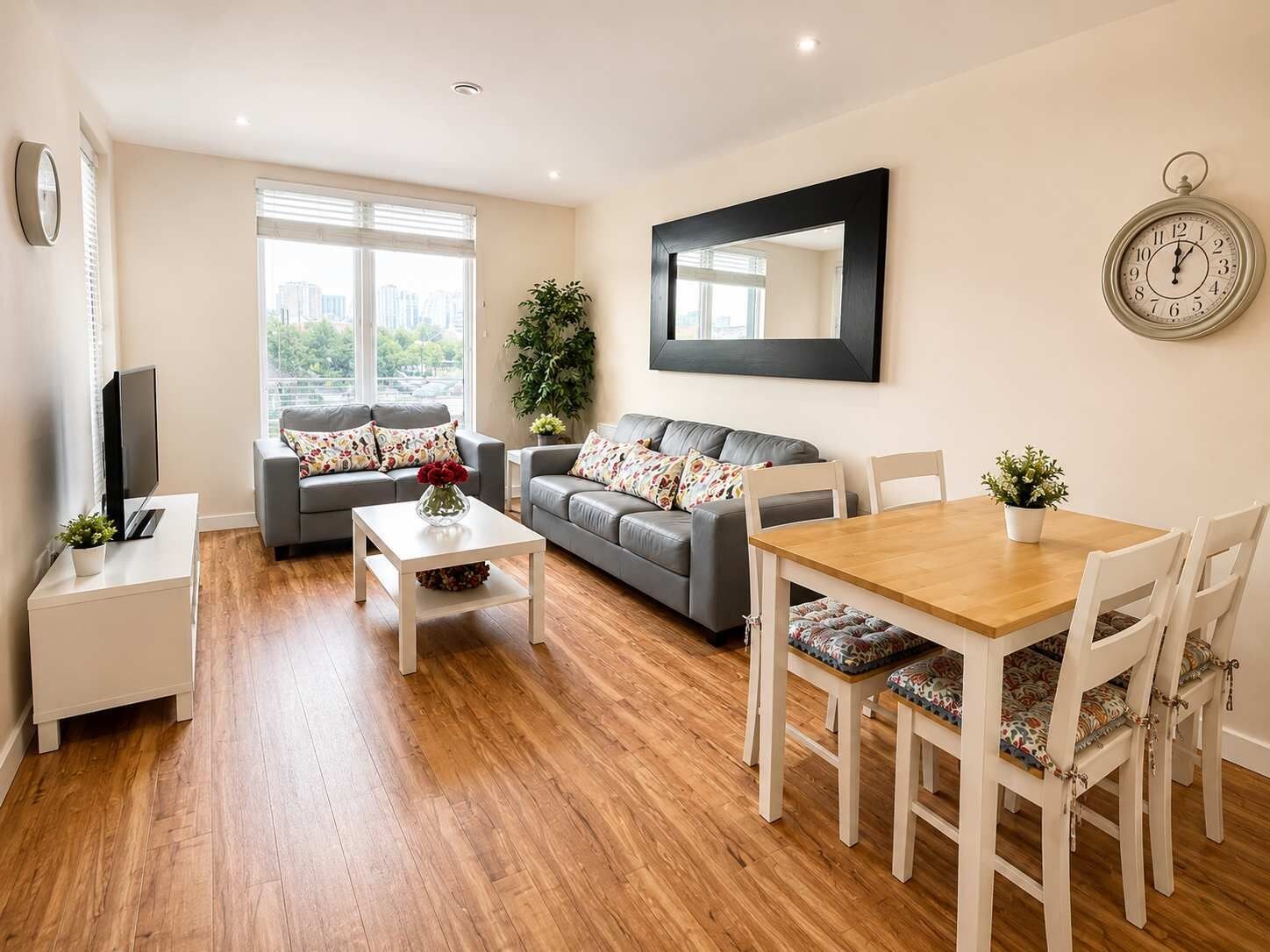 A modern living room with a sofa, a coffee table, a dining table with chairs, a TV, and a large window with blinds.