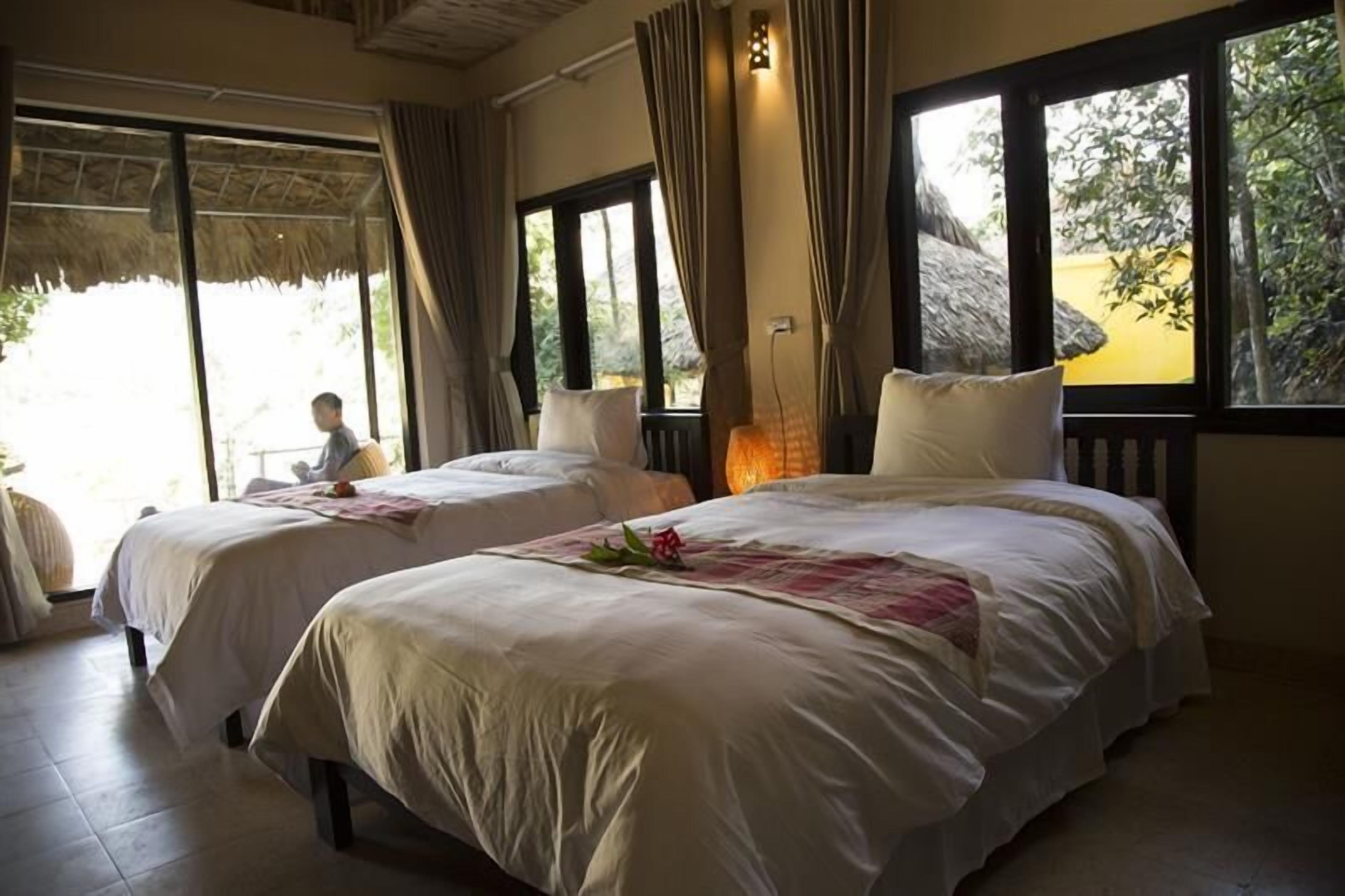 A room with two beds, large windows, and a view of greenery outside.