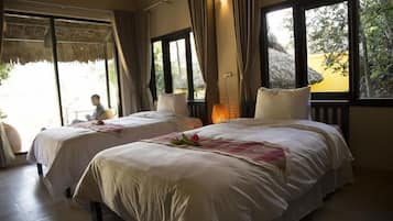 A room with two beds, large windows, and a view of greenery outside.
