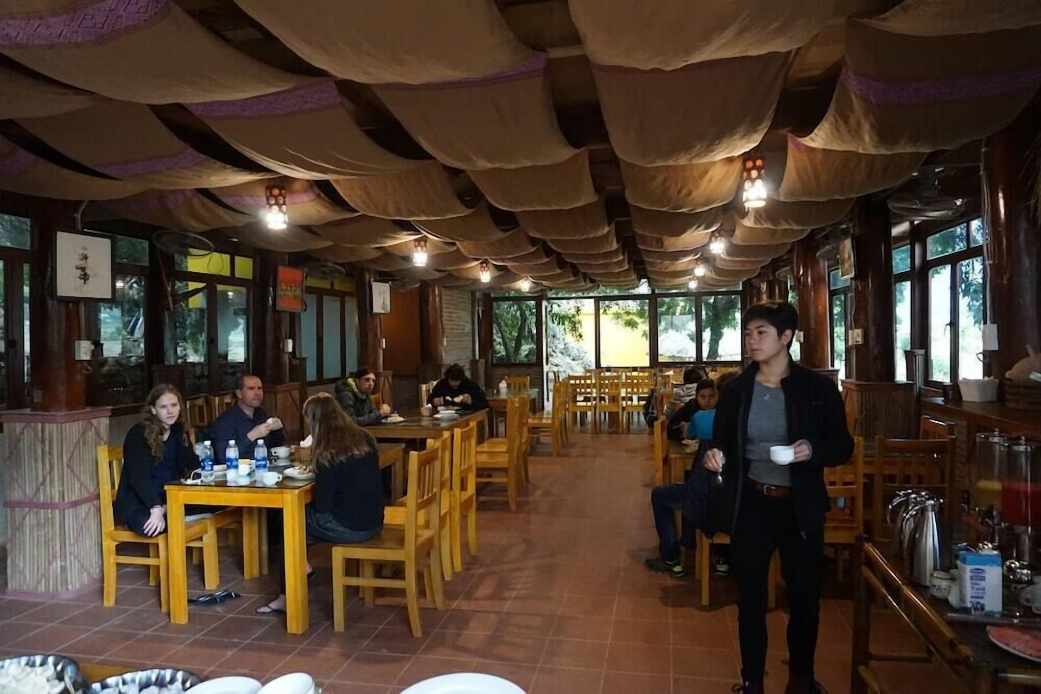 A restaurant with wooden tables and chairs, a high ceiling with hanging fabric, and large windows allowing natural light.