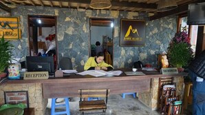 A reception desk with a receptionist, a 'RECEPTION' sign, and a 'SUNRISE HILLGATE' sign. There are two blue stools, a wooden chair, and a small table with a plant. - Mai Chau Nature Lodge - Hostel (Mai Chau)