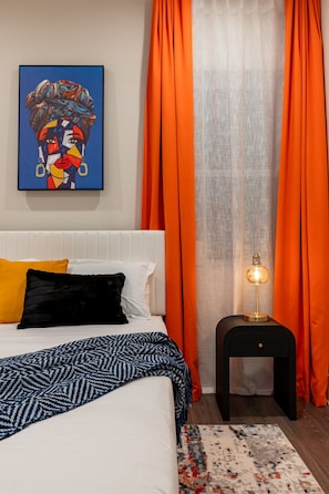 Ein Schlafzimmer mit einem Bett, Kissen, einem Nachttisch, einer Lampe, einem Gemälde und orangefarbenen Vorhängen.