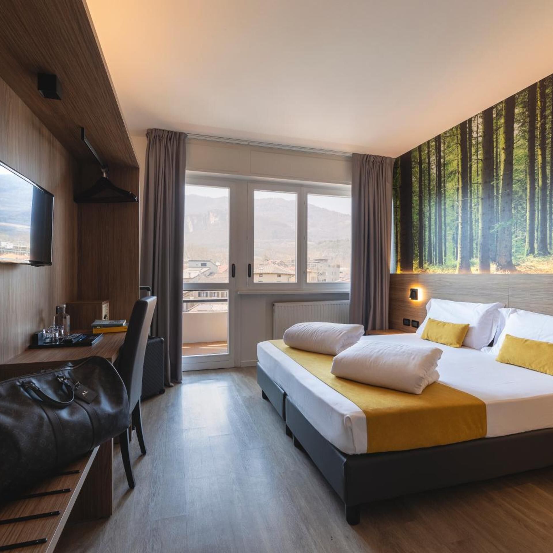A hotel room with a bed, a desk, a chair, a television, and a view of a forest through the window.