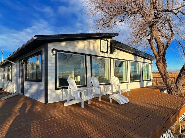 Una casa moderna con ventanas amplias, una terraza con sillas blancas y un árbol al fondo.
