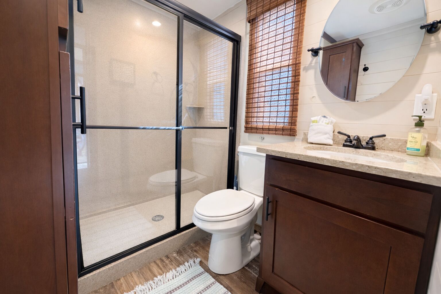 Une salle de bain avec une douche à parois de verre, des toilettes, un lavabo avec un miroir au-dessus et une fenêtre avec des stores.