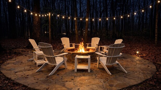 A circular stone fire pit with four Adirondack chairs around it, surrounded by a string of lights.