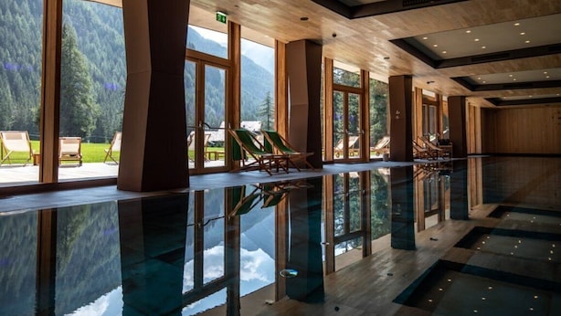 A spacious indoor swimming pool with large windows, wooden pillars, and a view of the outdoors. - Campzero Active & Luxury Resort (Ayas)