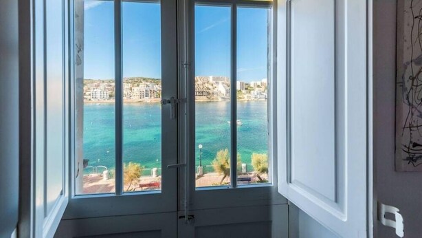 A room with a large window offering a view of a body of water and buildings across the water. - Blue Harbour Penthouse (St. Paul's Bay)