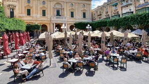 An outdoor cafe with tables and chairs, a large building in the background, and umbrellas providing shade. - Blue Harbour Penthouse (St. Paul's Bay)