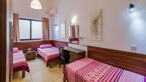 A hotel room with two single beds, a desk, a chair, a window with curtains, a wall-mounted air conditioner, a wall-mounted light fixture, and a framed artwork. - Blue Harbour Penthouse (St. Paul's Bay)