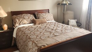 A neatly made bed with a patterned bedspread and pillows, a nightstand with a lamp, a chair, and a painting on the wall. - A cozy atmosphere and wonderful hosts make for a truly unforgettable experience. (Elliot Lake)