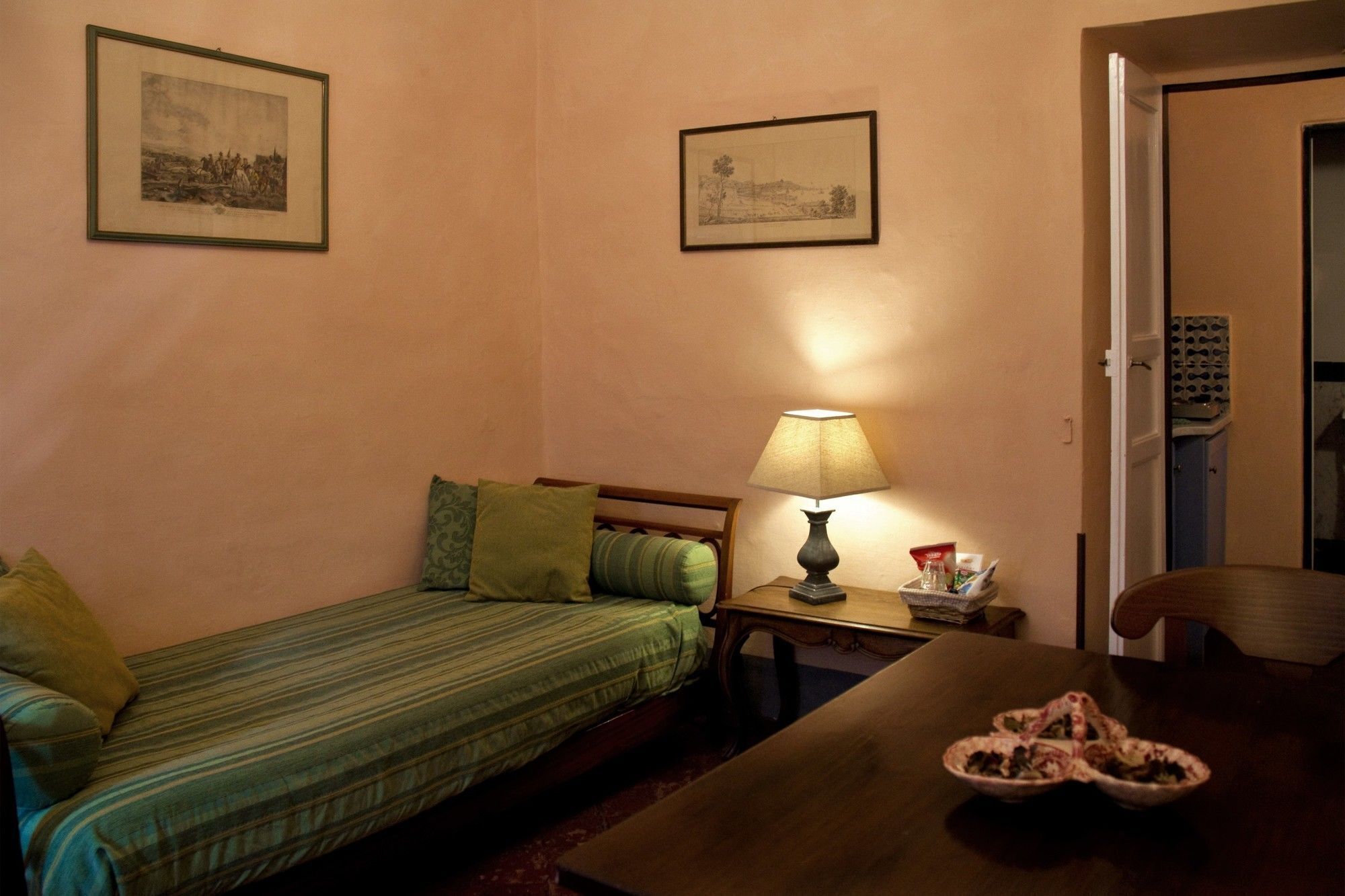 A small, cozy room with a single bed, a table with a lamp, and two framed pictures on the wall.