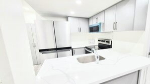 A modern kitchen with white cabinetry, a stainless steel refrigerator, a microwave, an oven, and a sink with a faucet. - Niagara Cozzy Suite (Thorold)