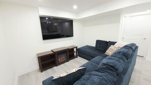 A modern living room with a large dark blue sofa, a flat-screen TV mounted on the wall, and a wooden entertainment unit with a fireplace. - Niagara Cozzy Suite (Thorold)