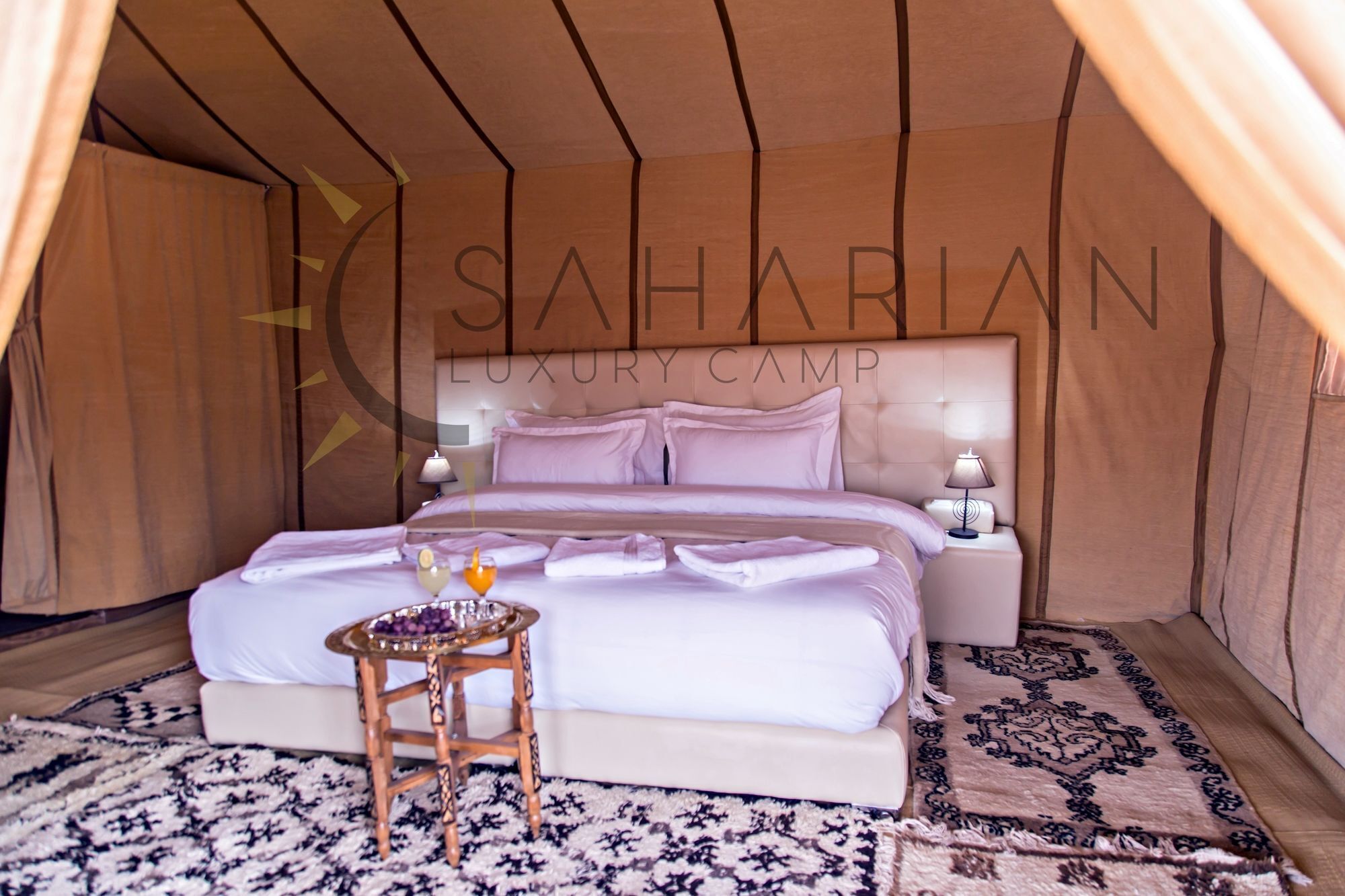 Quarto de um acampamento de luxo com tendas, com uma cama, abajures, uma mesinha com uma bebida e um tapete estampado.