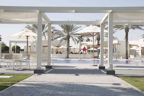 A pool area with white lounge chairs, umbrellas, and palm trees. - Sealine Beach, a Murwab Resort (Mesaieed)