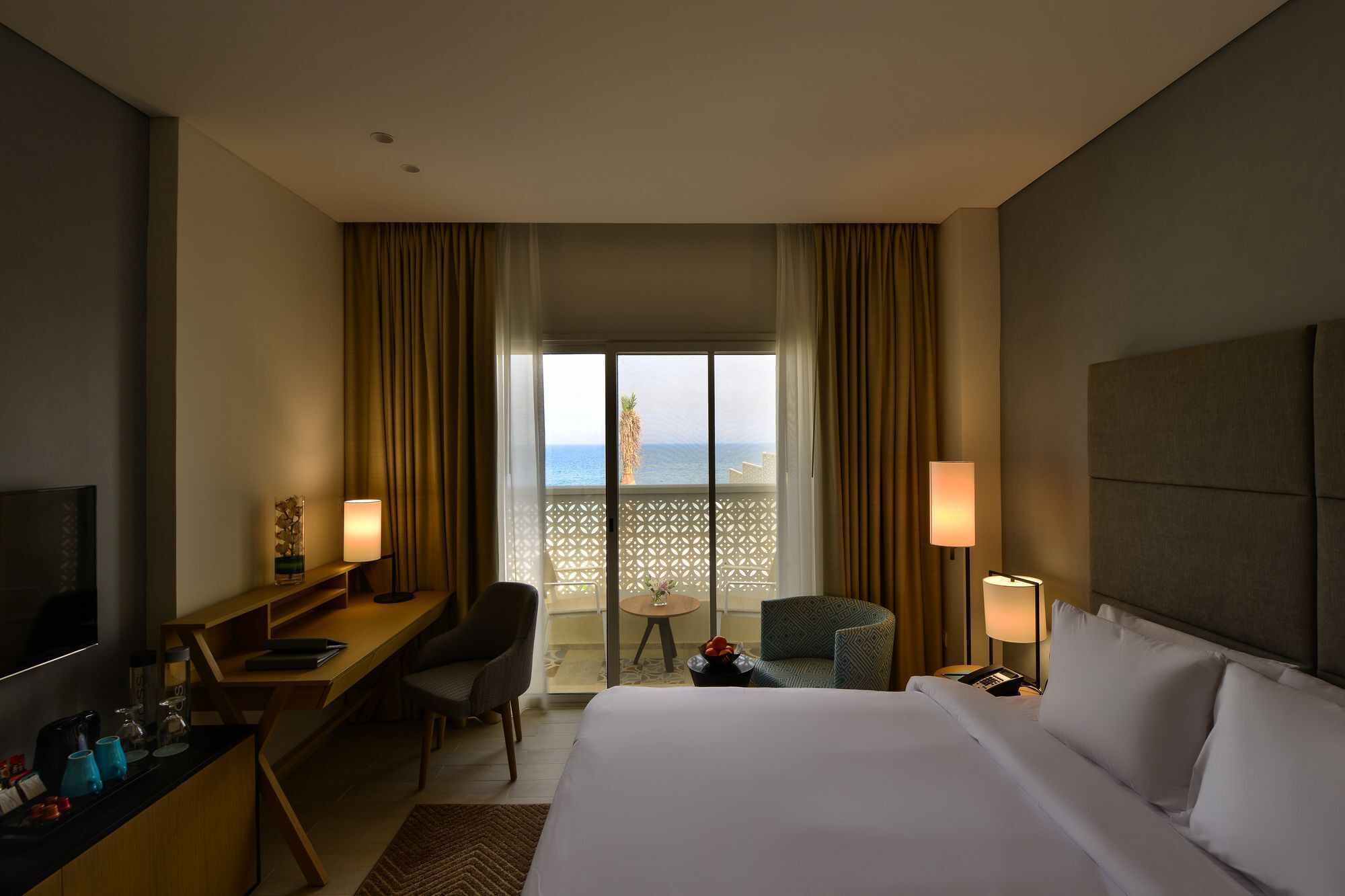 A hotel room with a bed, desk, chair, and a view of the sea.