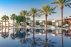 A pool area with lounge chairs and palm trees, surrounded by modern buildings. - Sealine Beach, a Murwab Resort (Mesaieed)