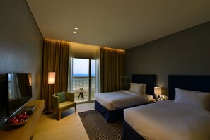 Ein Hotelzimmer mit zwei Betten, einem Fernseher, einem Schreibtisch mit Obstschale und Meerblick durch große Fenster.