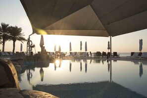 Un espace aménagé au bord de la piscine, avec des chaises longues et des parasols, une vue dégagée sur le coucher de soleil sur l’eau, et des palmiers en arrière-plan.