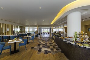 Un espace de restauration moderne dans un hôtel, avec de nombreuses tables et chaises, un buffet proposant une variété de plats, et une vue sur l’extérieur grâce à de grandes fenêtres.