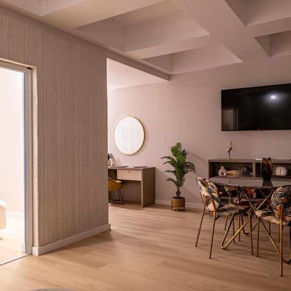 A modern living room with a flat-screen TV, a round mirror, a desk, and a dining area with chairs.