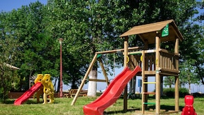 A playground with a wooden playhouse, a red slide, and yellow climbing structures. - Central Mamaia (Constanta)