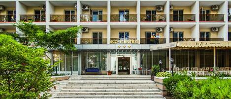 A multi-story hotel with a central entrance, outdoor seating area, and air conditioning units on the exterior.