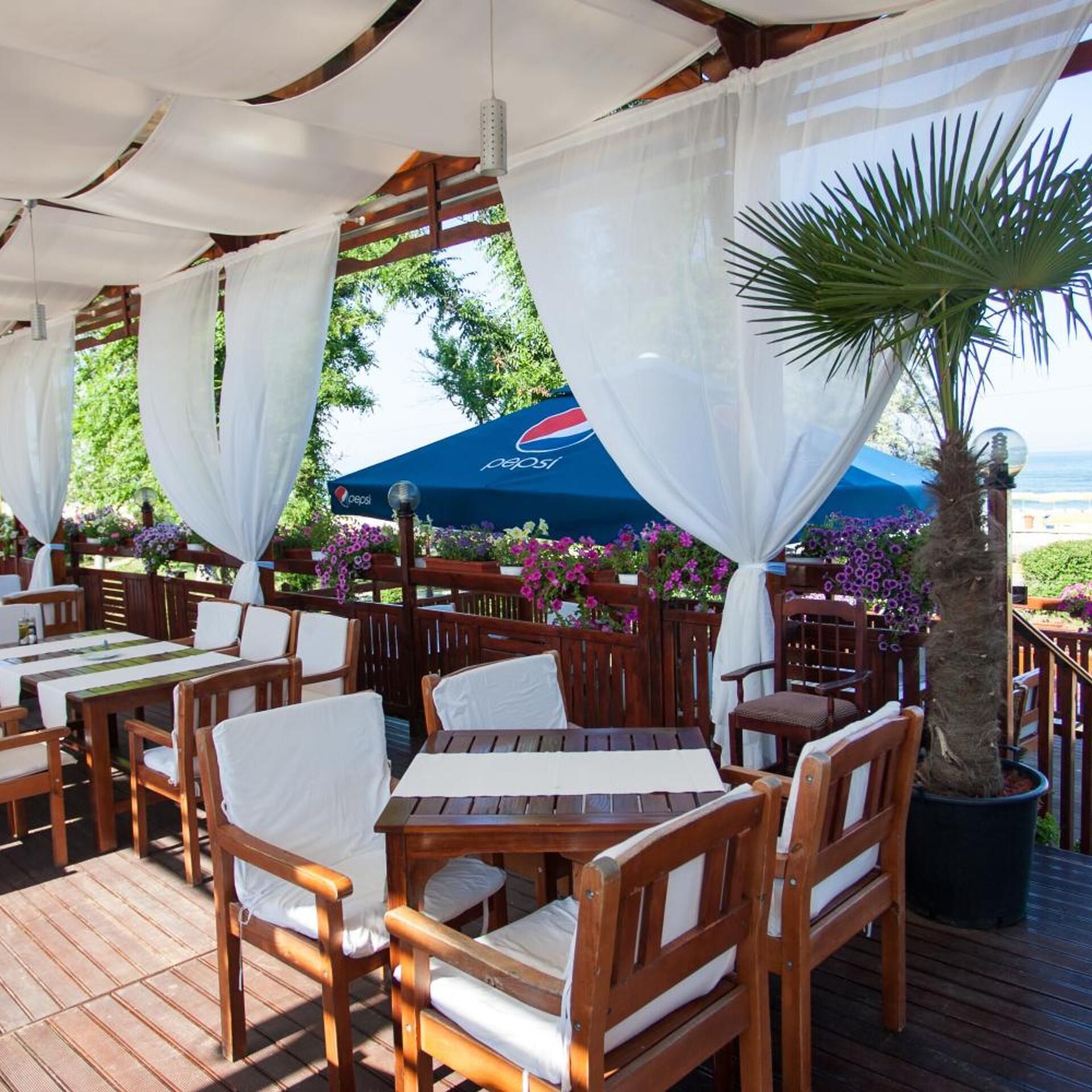 Un espace de restauration en plein air avec des tables et des chaises en bois, recouvert de rideaux blancs, et un parasol bleu orné du logo Pepsi.