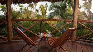 Une terrasse en bois, agrémentée de deux chaises en bois et d’une table ronde, entourée d’une balustrade en bois, et offrant une vue sur un jardin tropical avec des palmiers.
