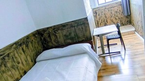 A single bed with a white sheet and a small table with a chair in a room with wooden flooring and a window with curtains.