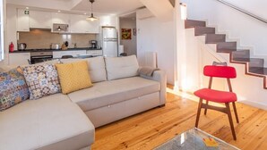 Una sala de estar moderna con un sofá, una mesa de centro de vidrio y una silla roja. Se ve una cocina con armarios blancos y un comedor al fondo.