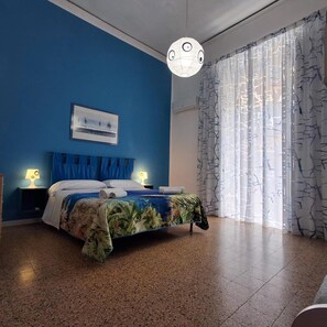 A bedroom with a bed, two bedside tables with lamps, a ceiling light fixture, a window with patterned curtains, and a wall-mounted air conditioning unit. - Mary''s Sweet Home Appartamento Napoli (Naples)