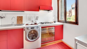Cocina compacta con armarios rojos, lavadora integrada y ventana con vistas a edificios.